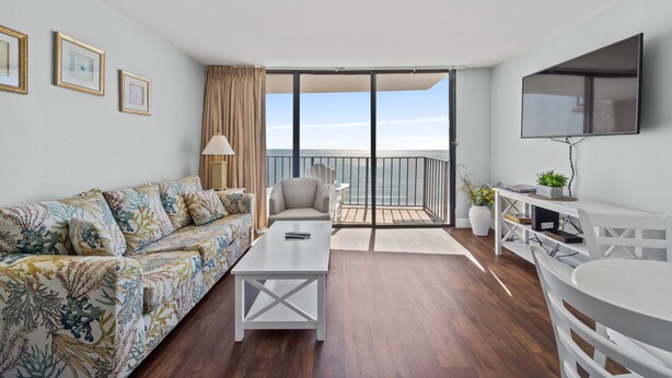 A modern living room with a sofa, a coffee table, a TV, and a balcony with a view of the sea. - Carolina Reef 804 - Ocean Front 1 Bedroom Unit - Great Views (North Myrtle Beach)
