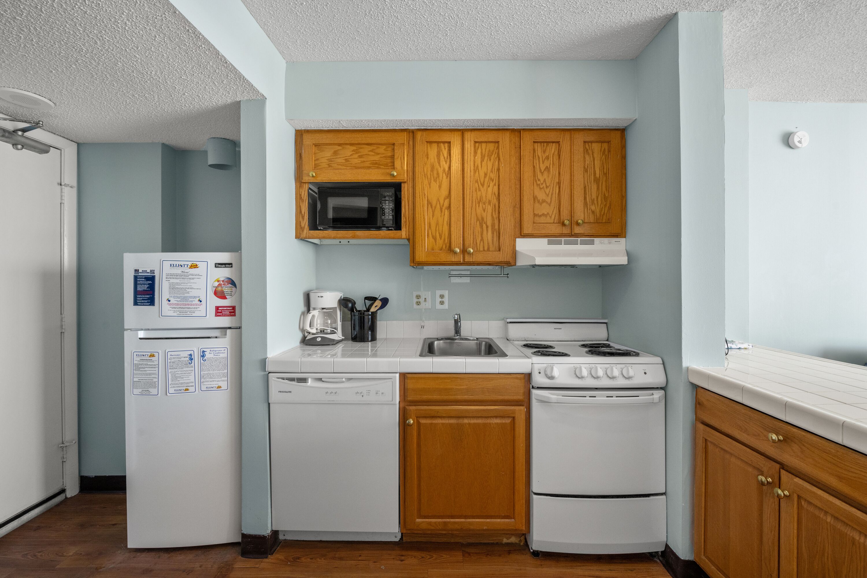 A compact kitchen with wooden cabinets, a microwave, a coffee maker, a dishwasher, and a stove.