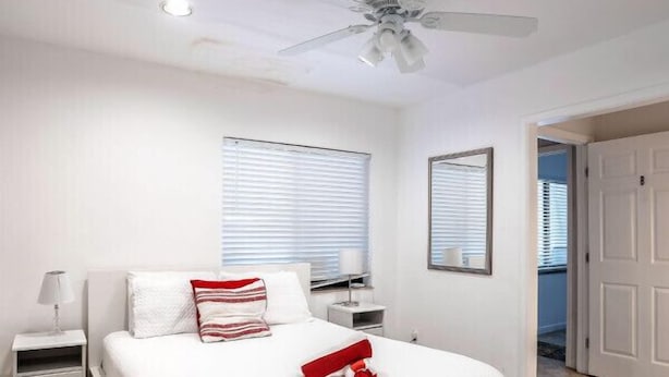 Une chambre moderne avec un grand lit, des murs blancs, un ventilateur de plafond et une fenêtre avec des stores.