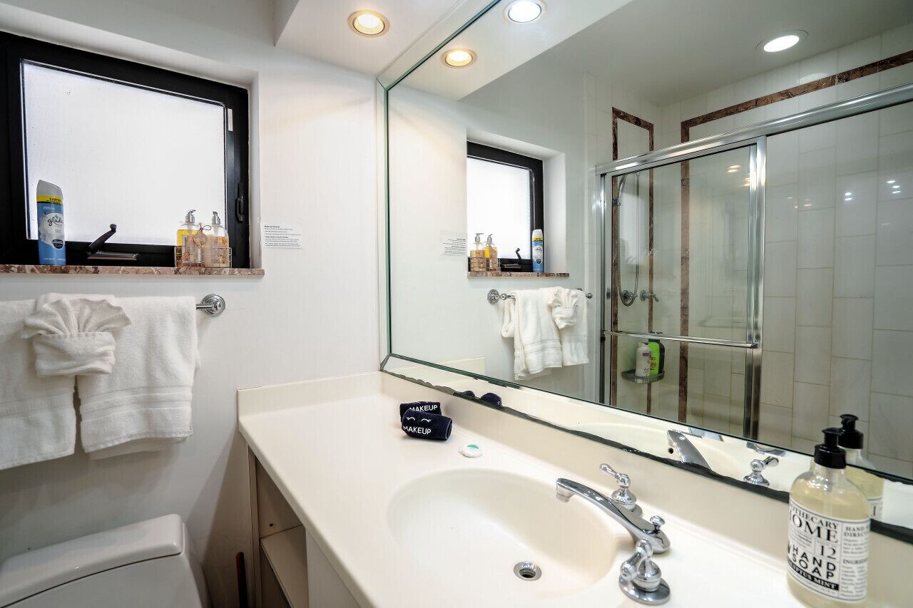 A bathroom with a large mirror, a sink, and a shower. There are towels hanging on the wall and toiletries on the counter.