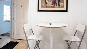 Une pièce avec une porte blanche, un tableau encadré représentant un couple en train de danser, une table ronde blanche et deux chaises blanches.