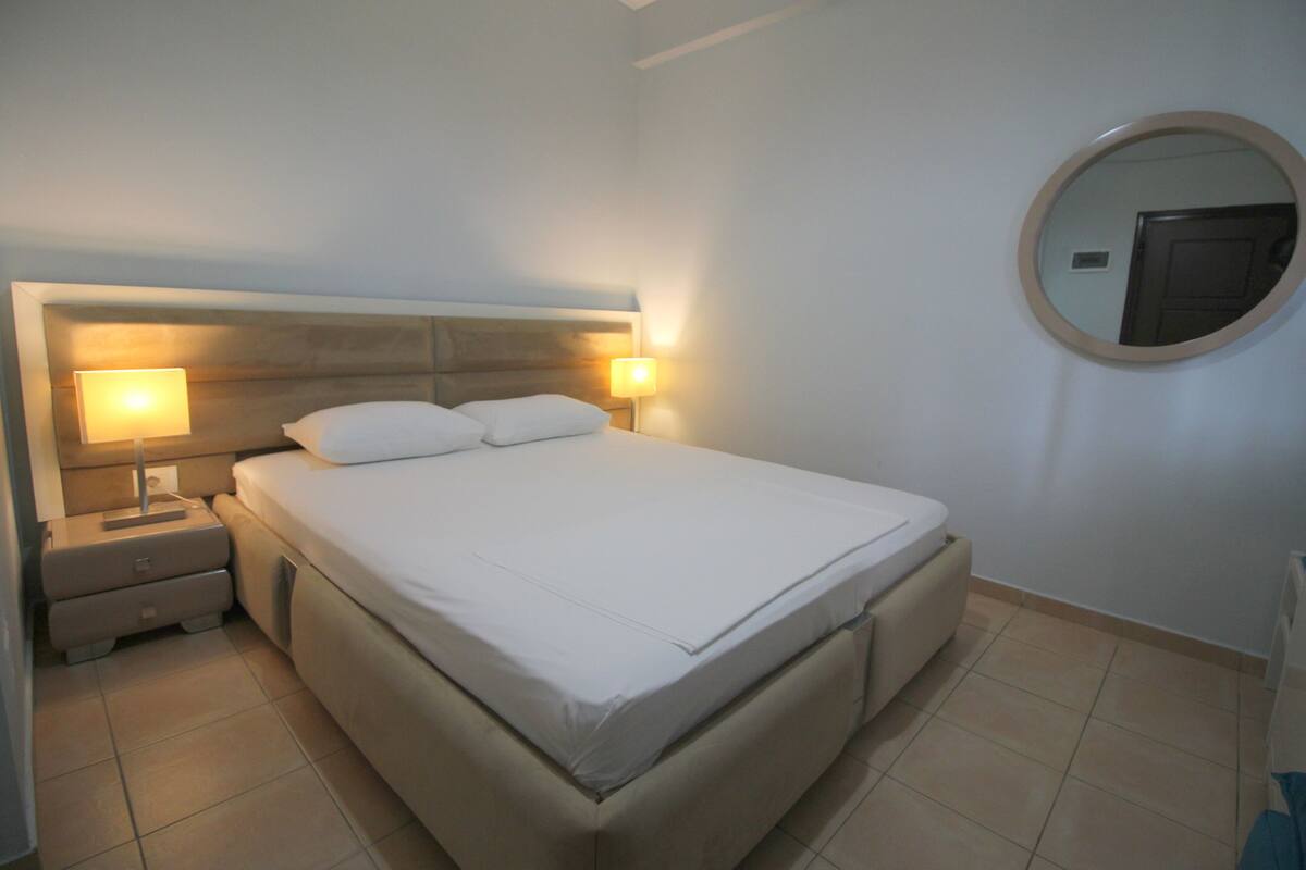 A neatly made bed with white linens, two bedside lamps, and a round mirror on the wall.