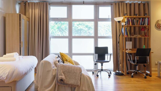 A hotel room with a bed, a desk, a chair, a bookshelf, and large windows. - Fantastically Located 2BDR Studio in Kennington (London)