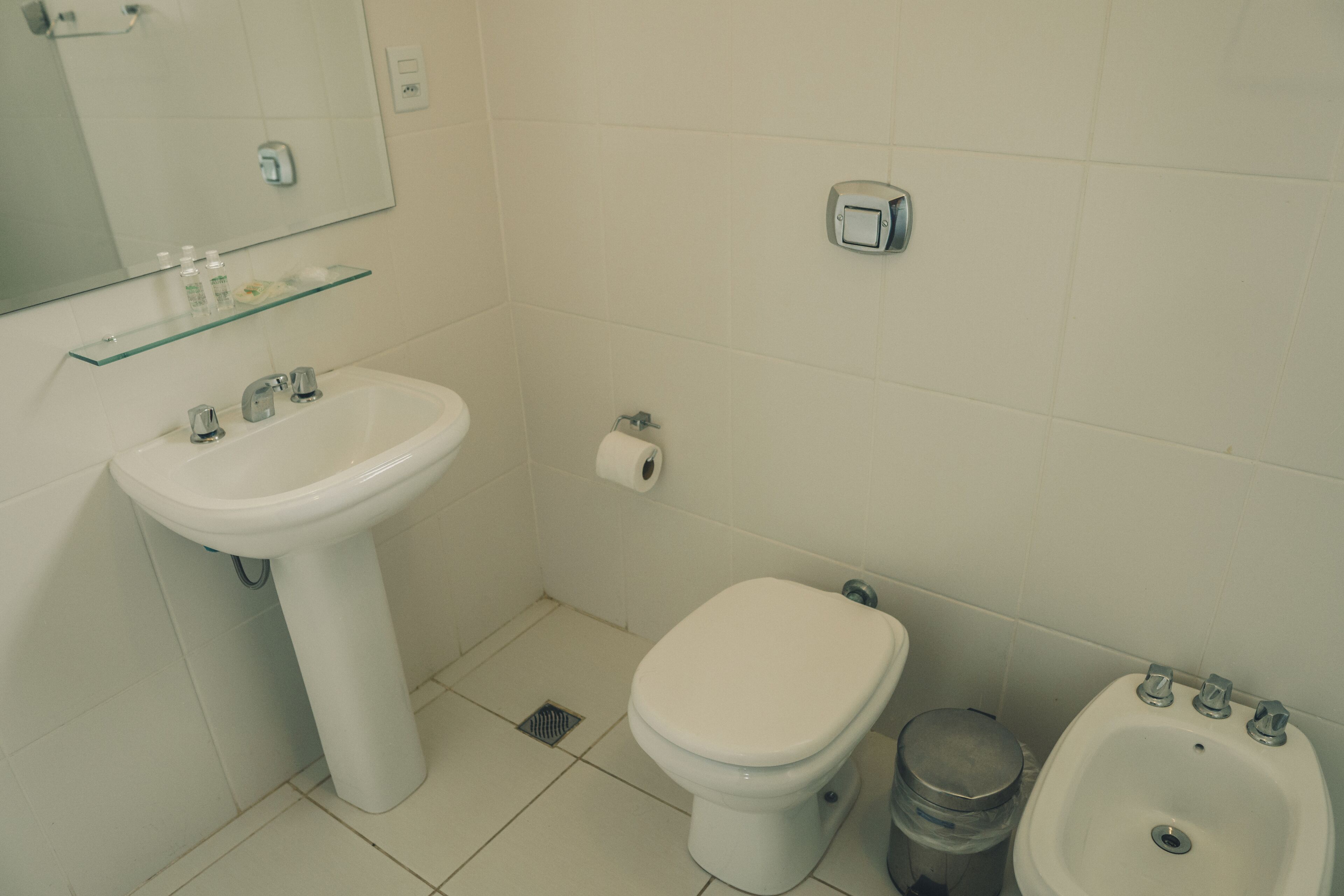 Un baño con lavamanos blanco, inodoro, bidé y estante de vidrio sobre el lavamanos.