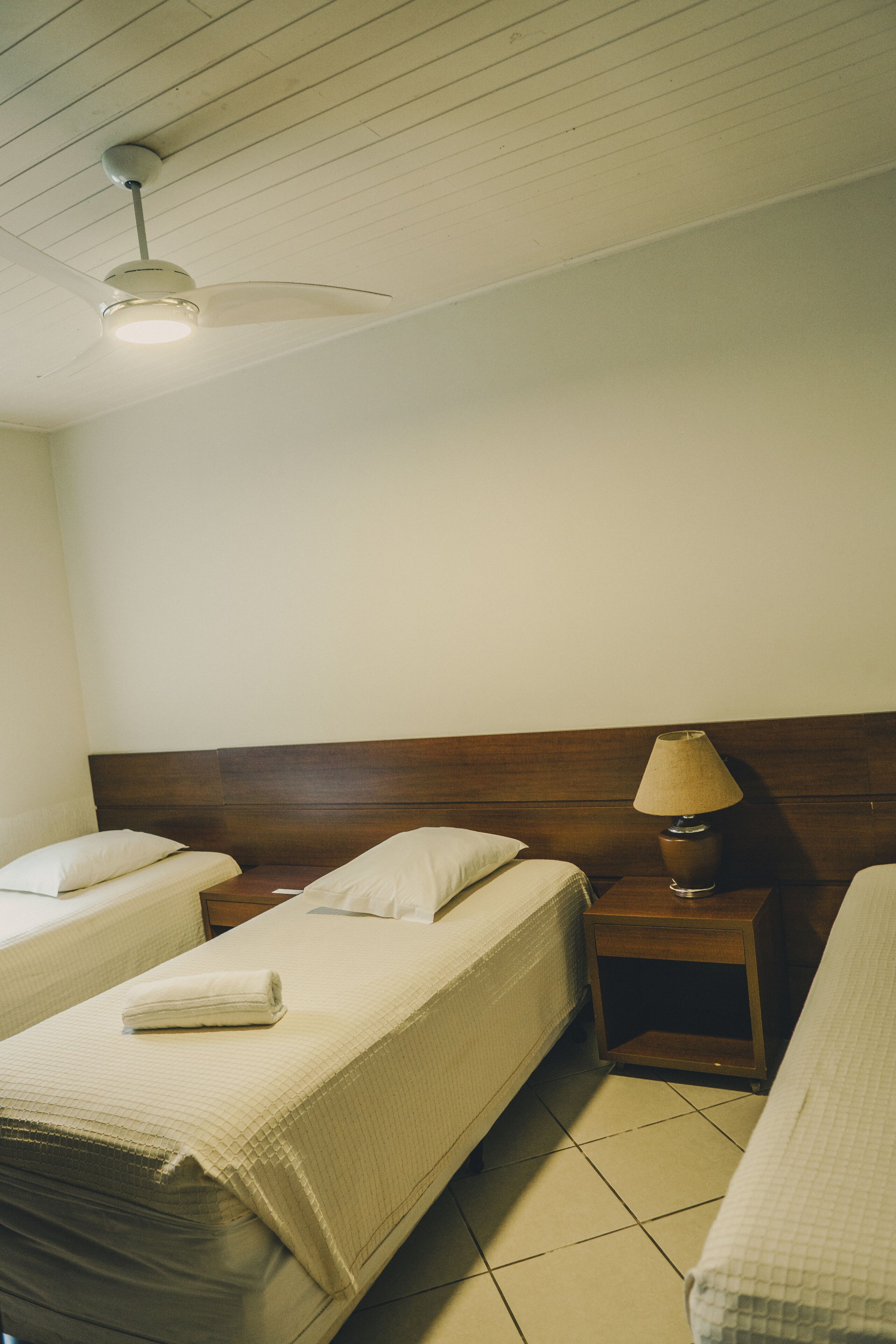 A hotel room with two single beds, a wooden headboard, a nightstand with a lamp, and a ceiling fan.