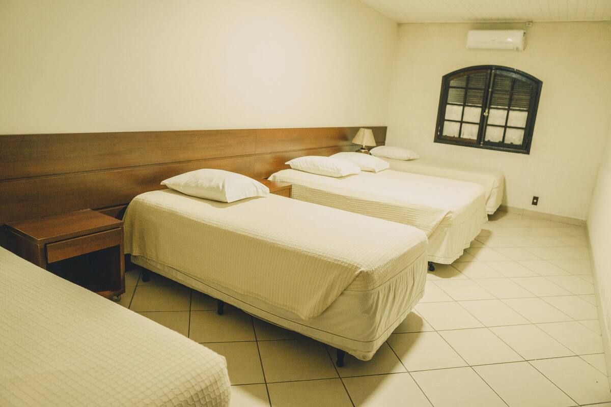 Ein Hotelzimmer mit drei Betten, einem Holzkopfende, einem Fenster und einem Fliesenboden.