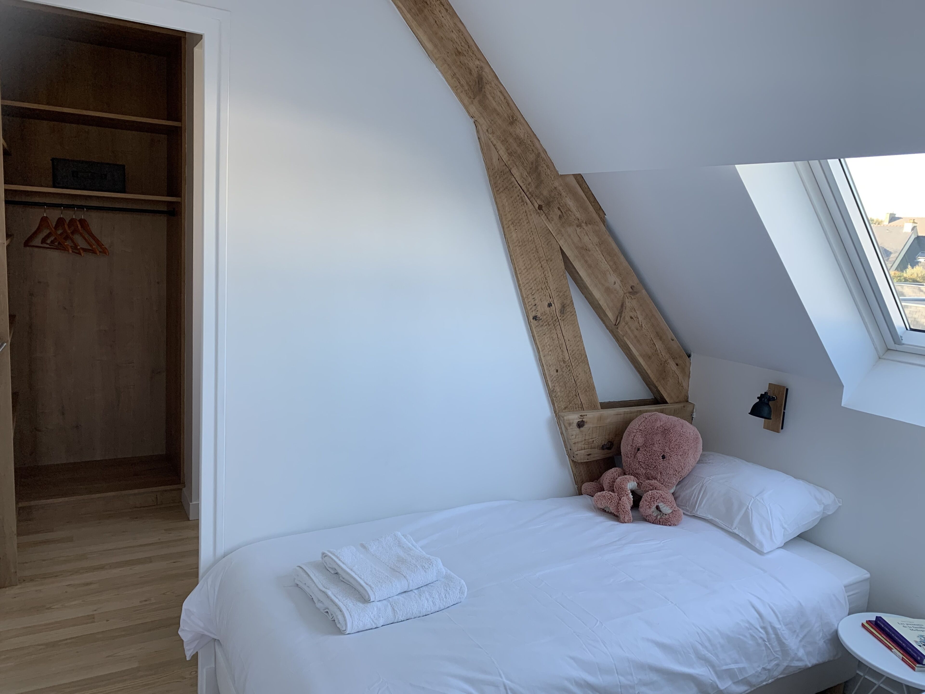 Een slaapkamer met een schuine plafond, een houten garderobe, een bed met witte lakens, een teddybeer en een klein nachtkastje met boeken.