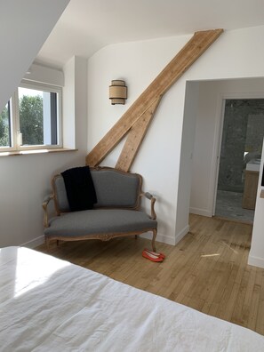A bedroom with a sloped ceiling, a wooden beam, a grey sofa with a black cushion, a white bed, and a wooden floor. - Maison Avec Piscine Intérieure Sainte-marine à 300m de la Plage (Combrit Sainte-Marine)