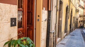 A narrow street with a wooden door and a potted plant on the sidewalk. - Arc Home - The charm of living in the heart of Castello, the oldest neighborhood. (Cagliari)