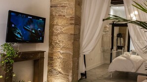 A room with a stone wall, a television mounted on the wall, a bed with white bedding, and a large plant in the foreground. - Arc Home - The charm of living in the heart of Castello, the oldest neighborhood. (Cagliari)