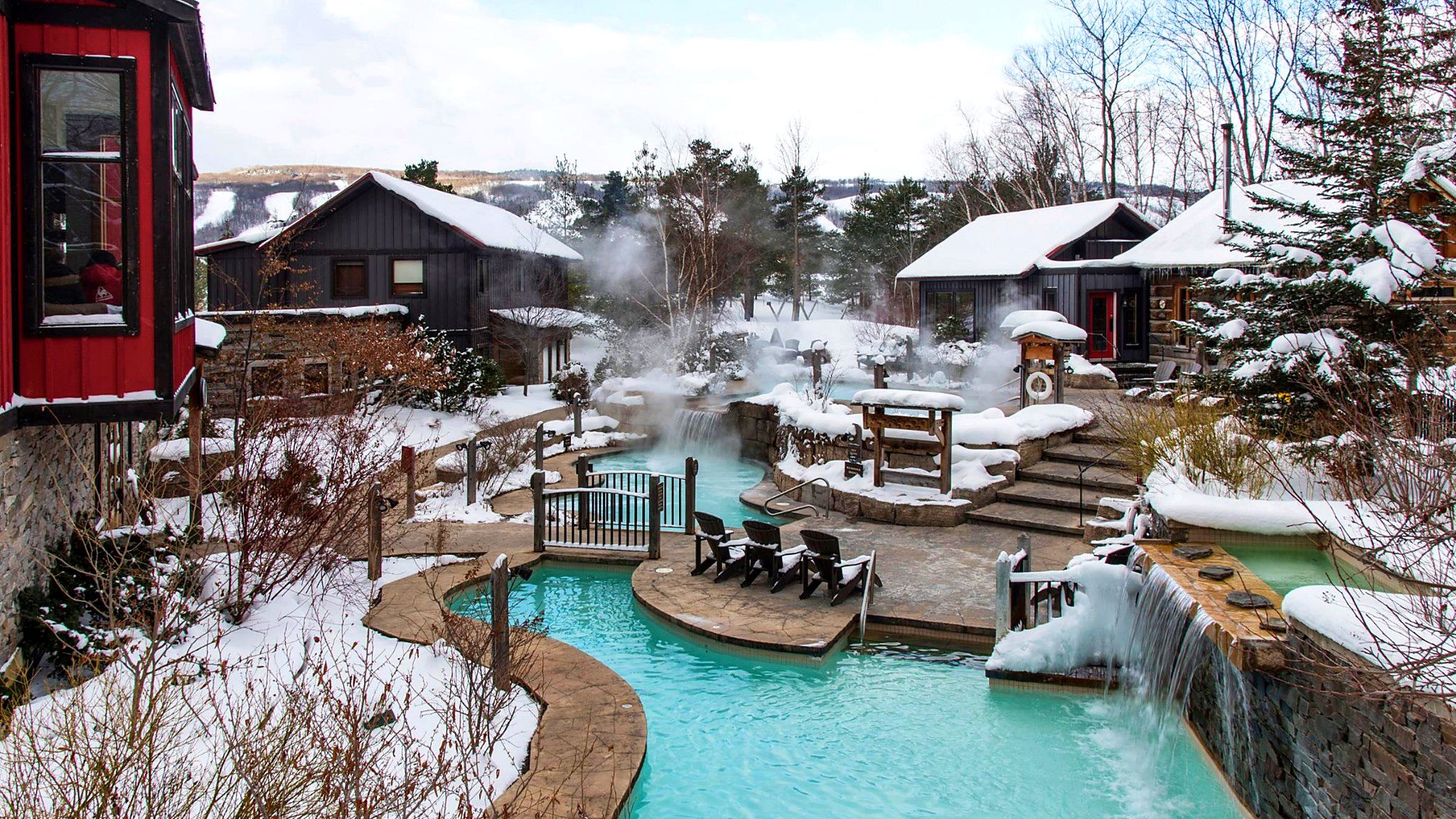 Una località turistica invernale con una piscina termale, poltroncine di legno e edifici ricoperti di neve.