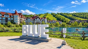 Un letrero con la palabra 'BLUE' en letras grandes, un lago con barcos y un fondo de casas y vegetación.