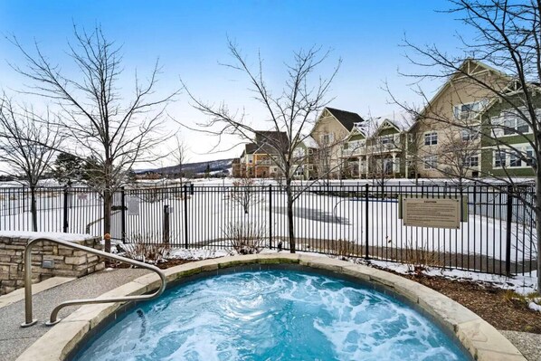 A swimming pool with a stone border, surrounded by a metal fence, with houses in the background.