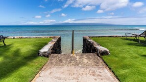 Eine Beton-Aussichtsplattform mit einem Metallpfosten in der Mitte, mit Blick auf einen Strand und klares blaues Wasser sowie eine entfernte Insel.