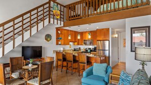 Ein geräumiger Wohnbereich mit einem blauen Sofa, einem Holzespeisetisch und Stühlen. Man sieht eine Küche mit Holzausstattung und einem Edelstahlkühlschrank. Eine Holztreppe führt auf eine obere Ebene.