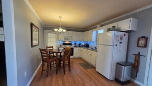 Une cuisine avec un plancher en bois, des armoires blanches, une table à manger avec des chaises, un réfrig érateur et une poubelle.