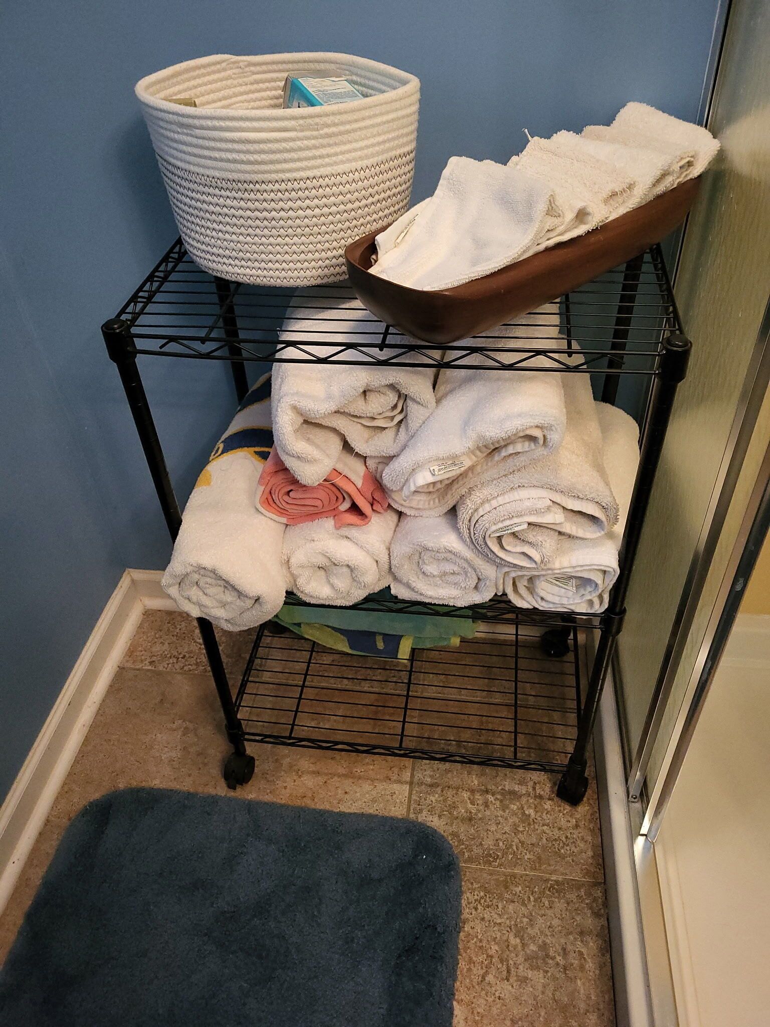 A black metal rack with folded towels, a white woven basket, and a brown bowl.