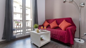 A modern living room with a red sofa, a white side table with a tray, a floor lamp, and a view of buildings through large windows. - New apartment in the Center, Eixample Best Area! 2 minutes to Passeig de Gracia  (Barcelona)