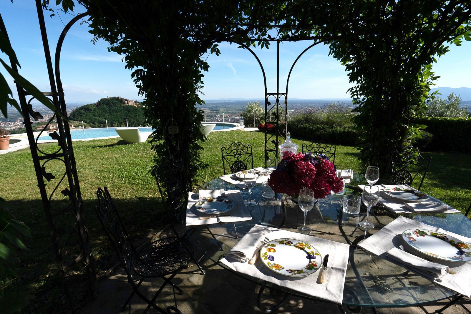 Un área de comedor al aire libre con mesas dispuestas para dos personas, rodeada de follaje y vistas a un paisaje urbano.