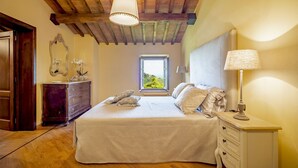 Ein Schlafzimmer mit Holzbalkendecke, einem großen Bett mit weißer Bettwäsche, einem Schminktisch, einem Spiegel und einem Fenster mit Blick auf Grünflächen.