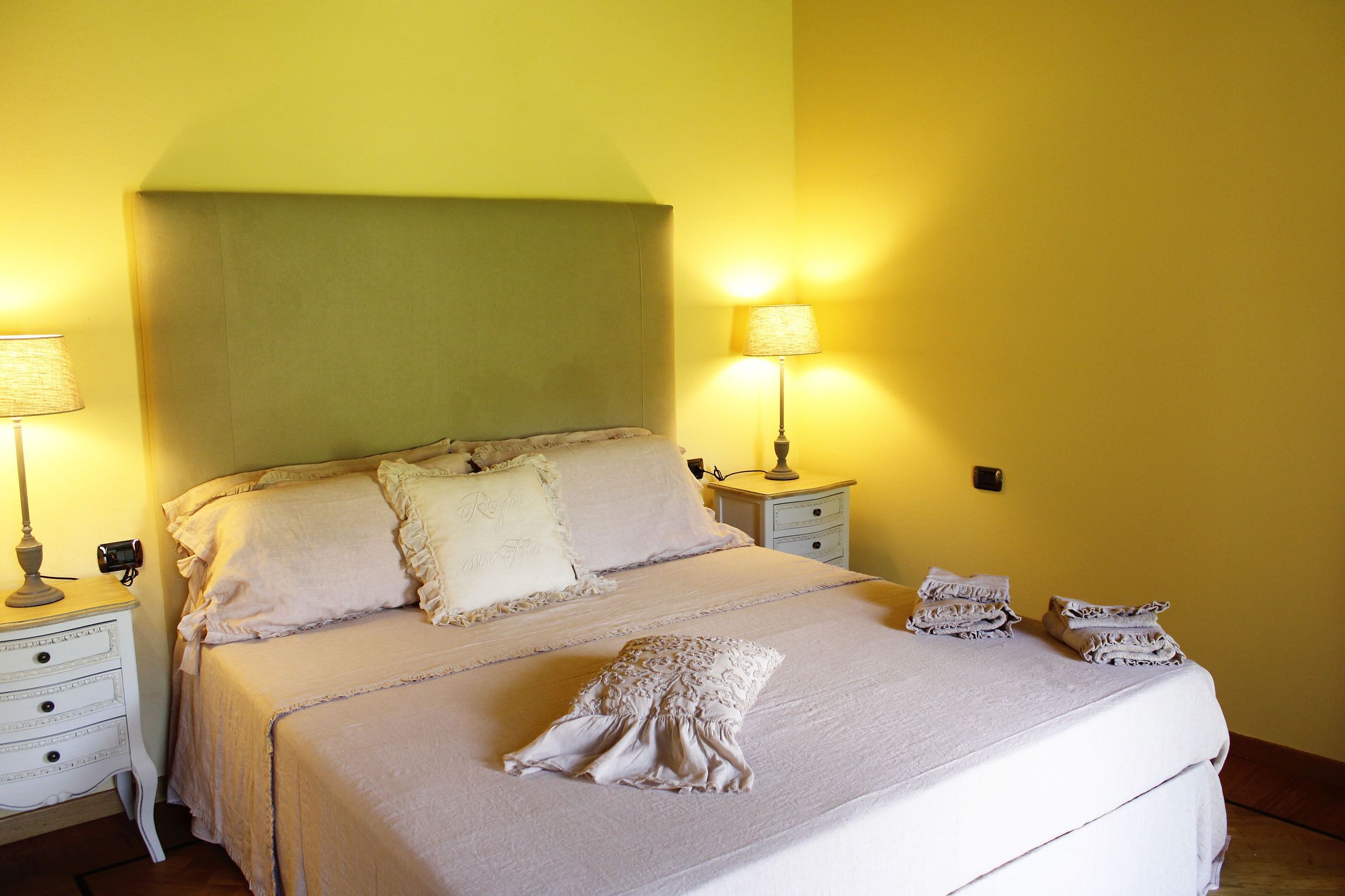 A neatly made bed with pillows and a folded towel on a white bedspread. There are two bedside tables with lamps and a small drawer.