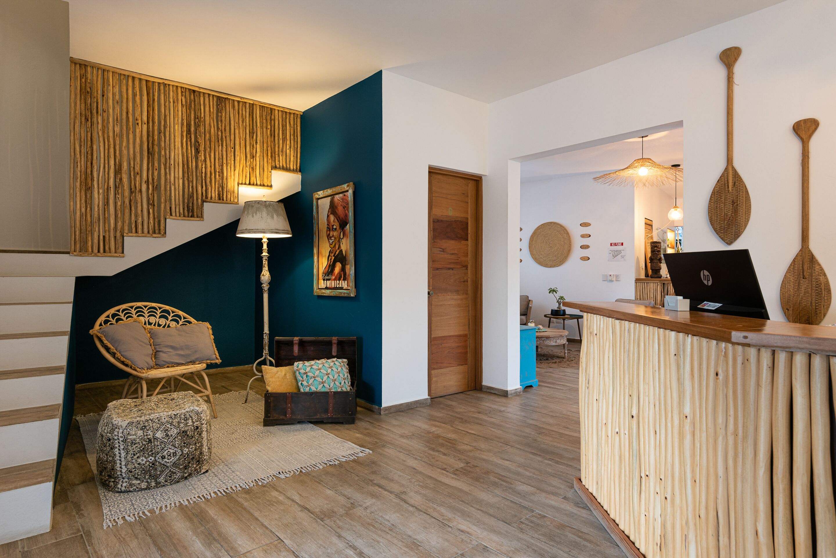 Un área de recepción con un escritorio de madera, una silla de mimbre, un reposapiés estampado, una escalera con pared azul y una puerta de madera.