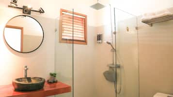 Un baño con un espejo redondo, un mueble de tocador de madera, una ducha con cerramiento de vidrio y un inodoro.