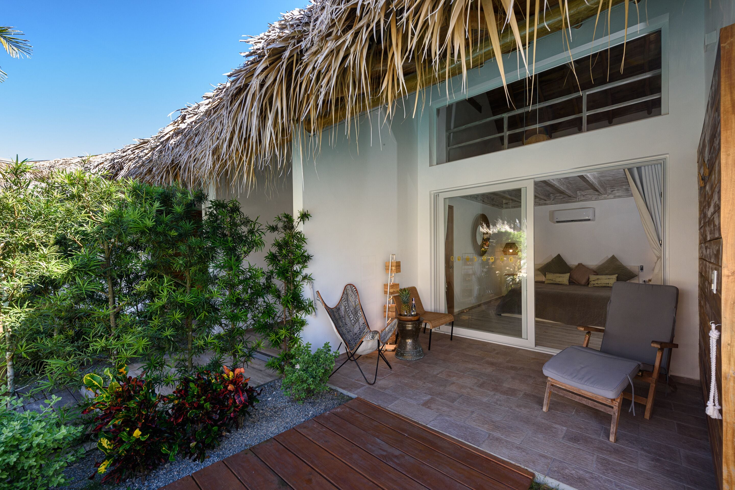 Una villa moderna con techo de paja, terraza de madera y puerta de cristal que da a un dormitorio con cama y almohadas.