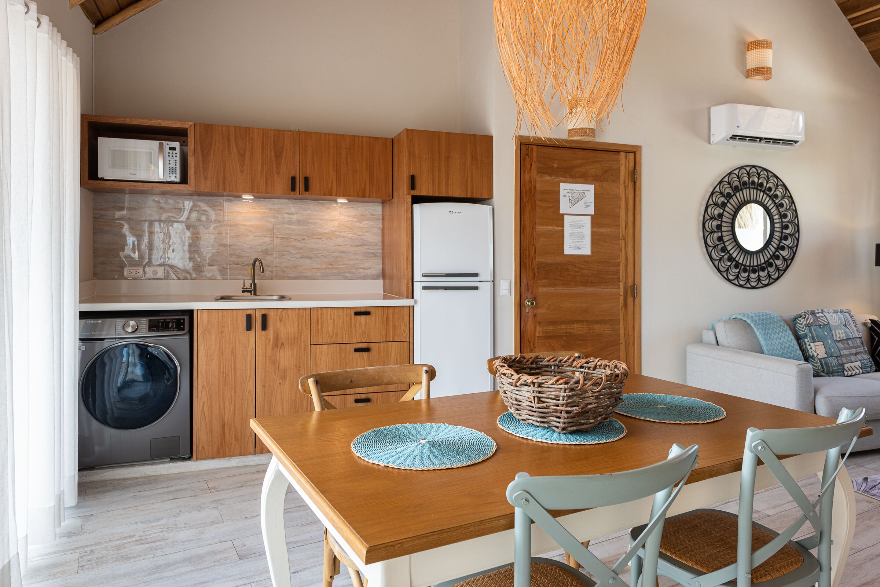 A modern kitchen with wooden cabinets, a built-in microwave, and a refrigerator. There's a dining table with blue placemats and a basket.