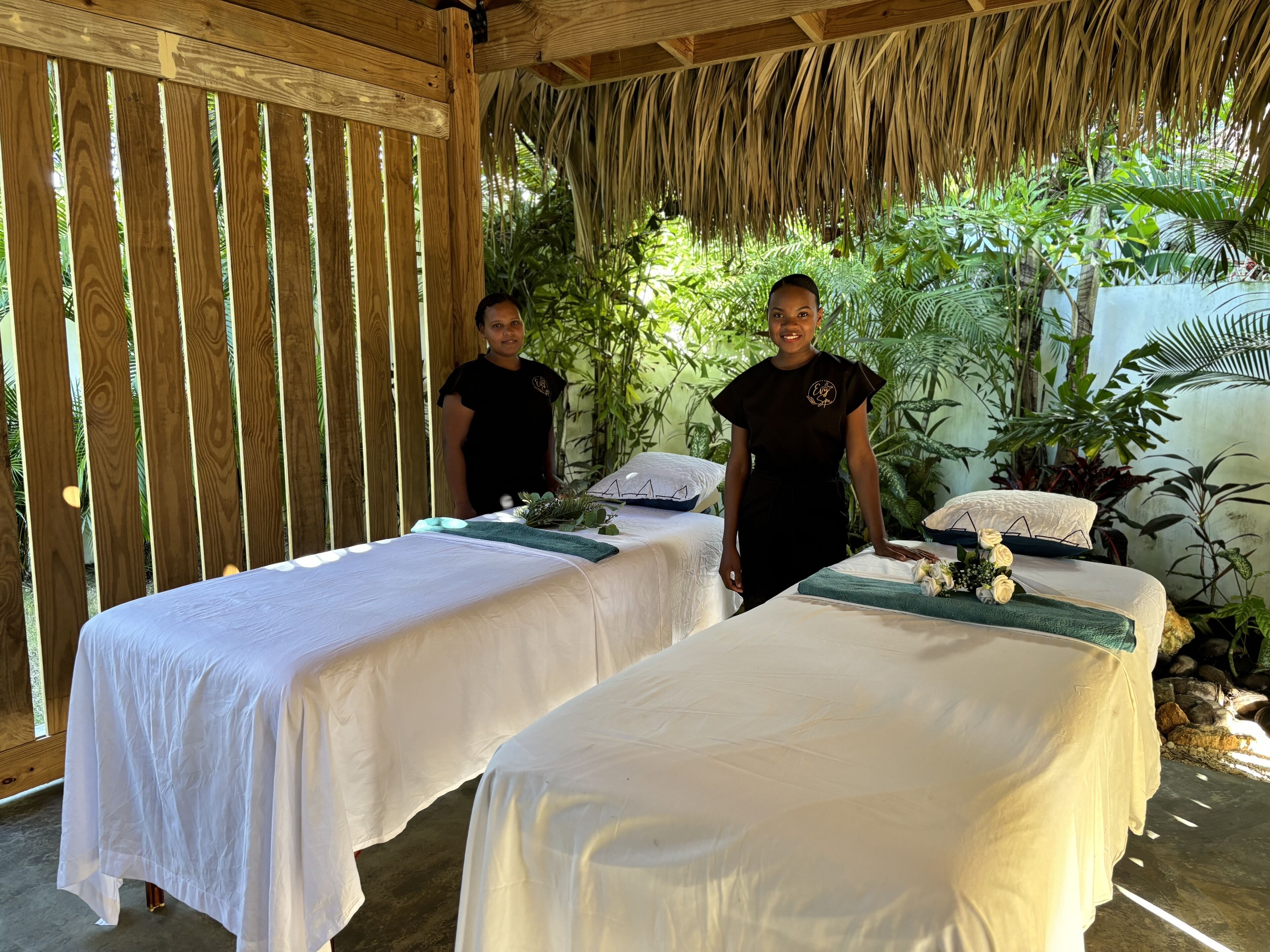 Two massage tables with white linens, a wooden partition, and lush greenery in the background.