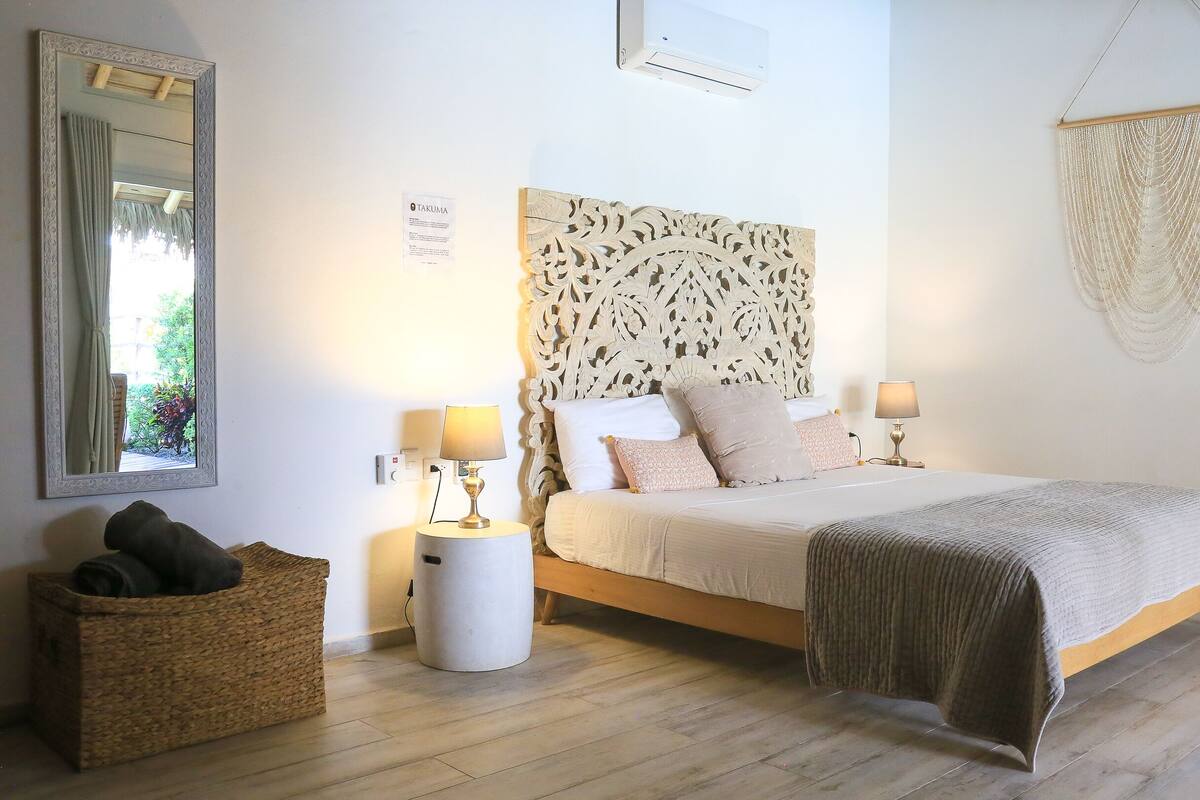 A bedroom with a bed, two bedside tables with lamps, a large mirror, a wall-mounted air conditioner, and a woven basket.