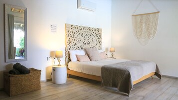 Un dormitorio con una cama, dos mesitas de noche con lámparas, un espejo grande, un aire acondicionado de pared y una cesta tejida.