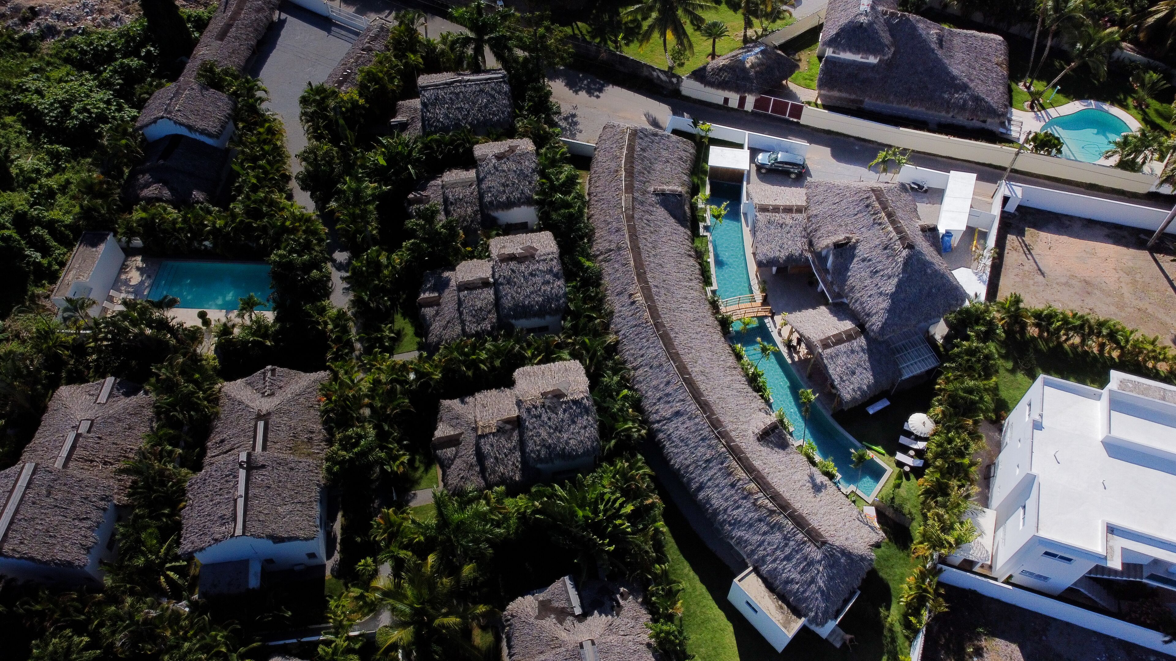 An aerial view of a resort with thatched-roof buildings, a central swimming pool, and lush greenery.
