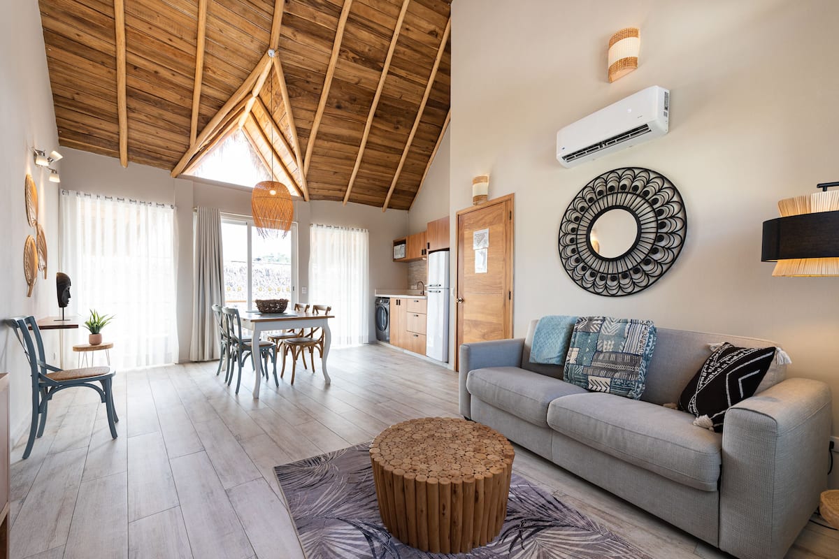 A spacious living area with a wooden ceiling, a grey sofa, a round wooden coffee table, and a dining area with a table and chairs.