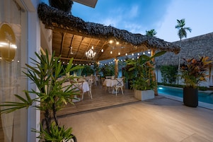 Un espacio de comedor al aire libre con mesas y sillas de madera, techo de paja y una piscina al fondo.