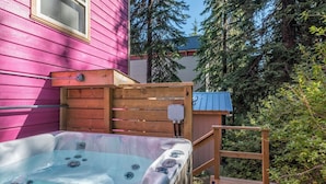 A hot tub adjacent to a pink house, surrounded by a wooden deck and dense greenery. - Family Orientated Lower Knoll Chalet All to Yourself w/Hot Tub, Laundry, BBQ (Silver Star Mountain)