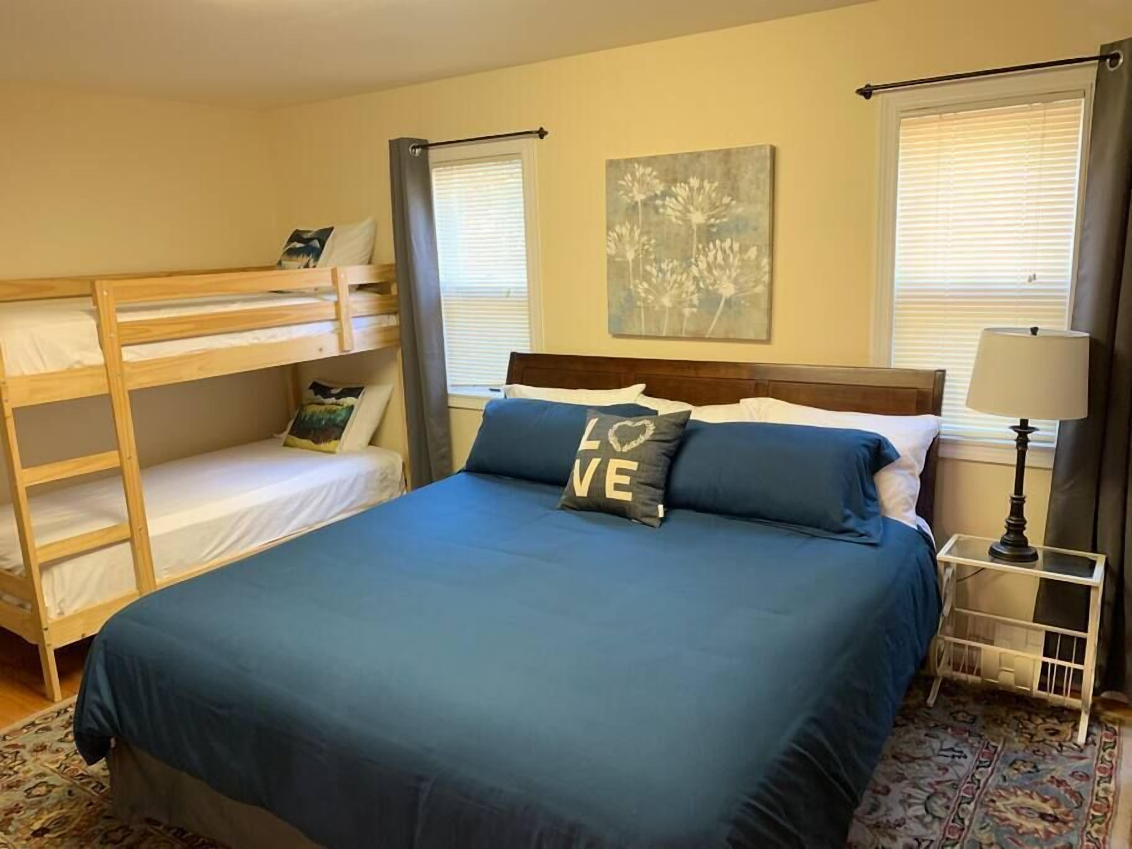 A bedroom with a bunk bed, a single bed with blue bedding, a nightstand with a lamp, and a floral painting on the wall.