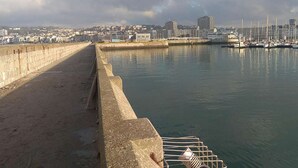 Eine Uferpromenade mit Betonbarriere, ruhigem Wasser und einem Jachthafen mit Booten.