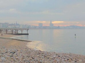 Une plage de galets avec une jetée s’avançant dans des eaux calmes, une vue sur les gratte-ciel de la ville en arrière-plan, et une personne pratiquant le paddle.