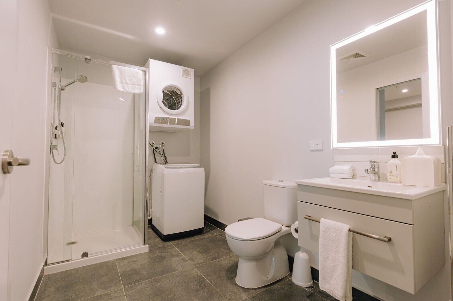 Baño con ducha de vidrio, inodoro, lavadora y tocador con espejo.