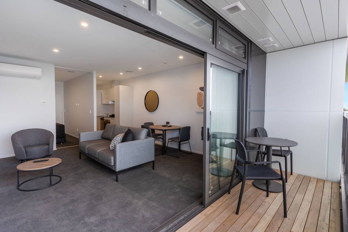 Un salon moderne avec un canapé gris, des fauteuils, une petite table ronde et une porte vitrée donnant sur un espace extérieur doté d’une terrasse en bois.
