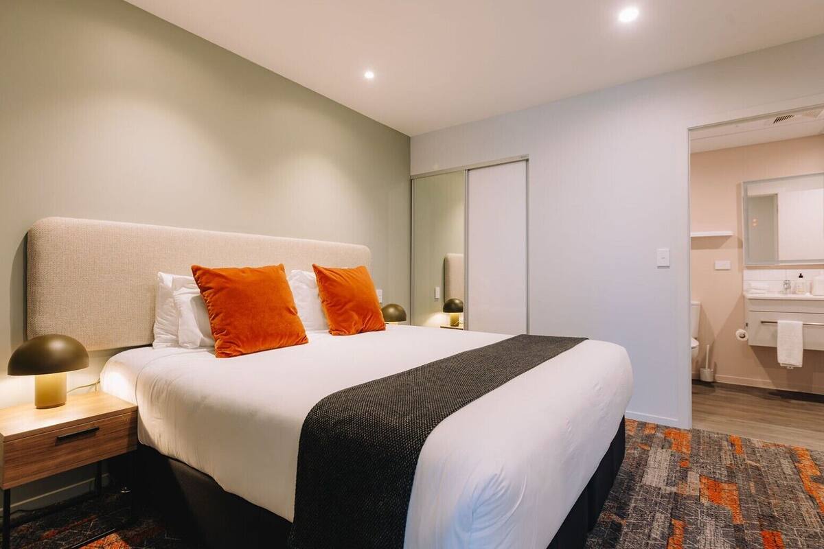 Une chambre d’hôtel avec un lit, une table de chevet, une lampe et une salle de bain visible à travers une porte ouverte.