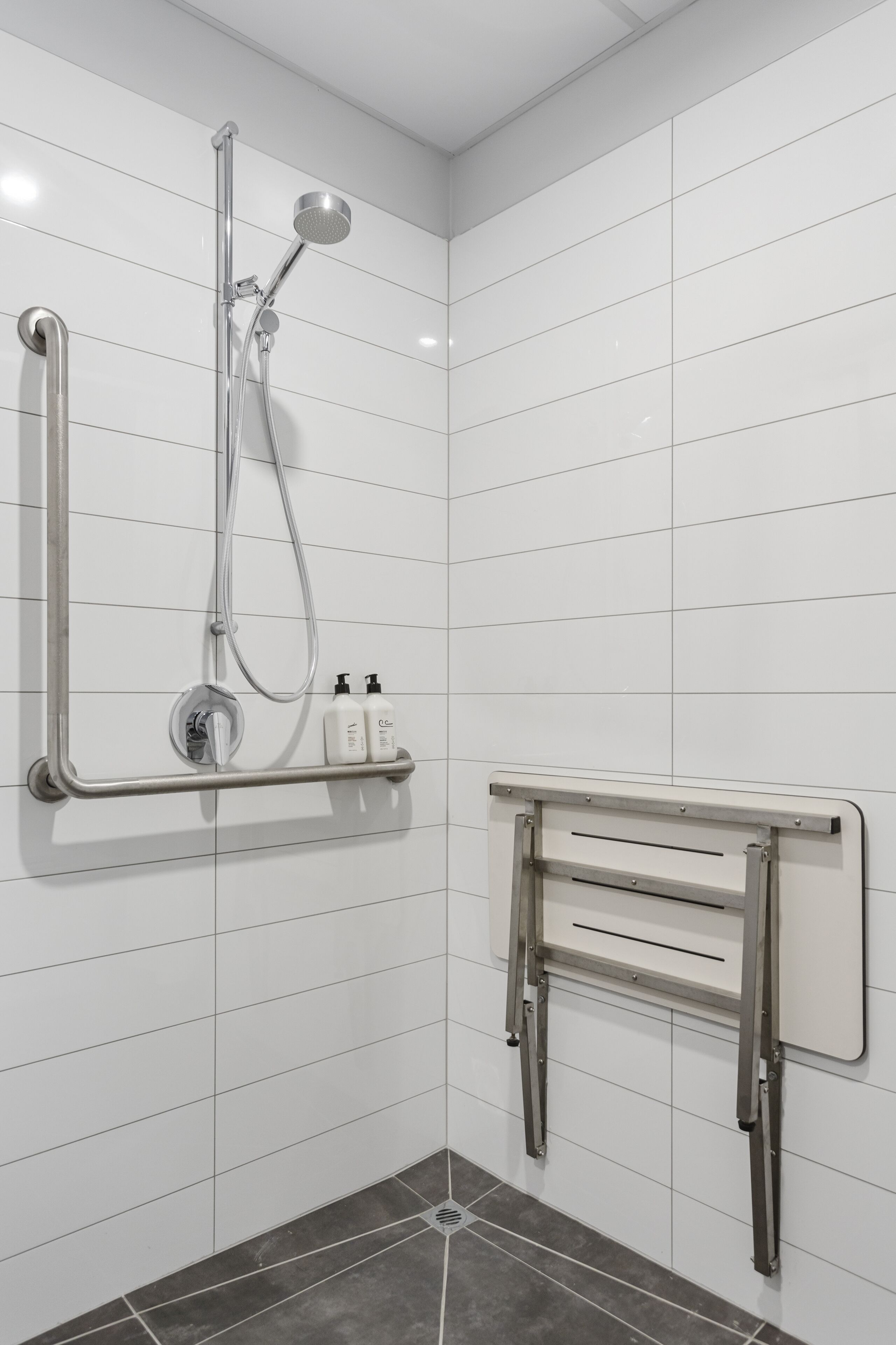 A modern bathroom with white tiled walls, a shower with a handheld showerhead, and a grab bar.