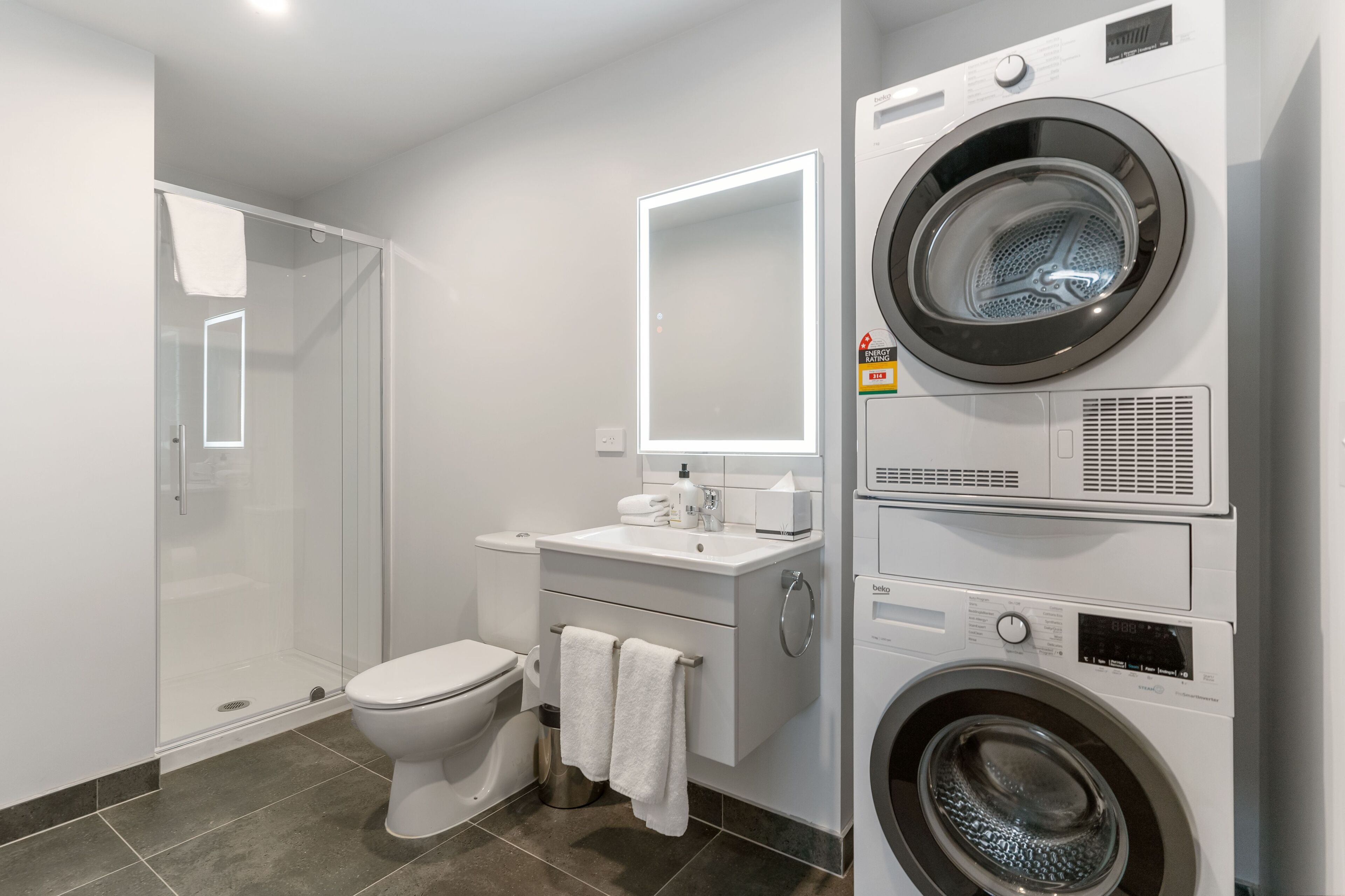 Un baño moderno con ducha envuelta en vidrio, un inodoro, un lavamanos con espejo y una lavadora y secadora apiladas.