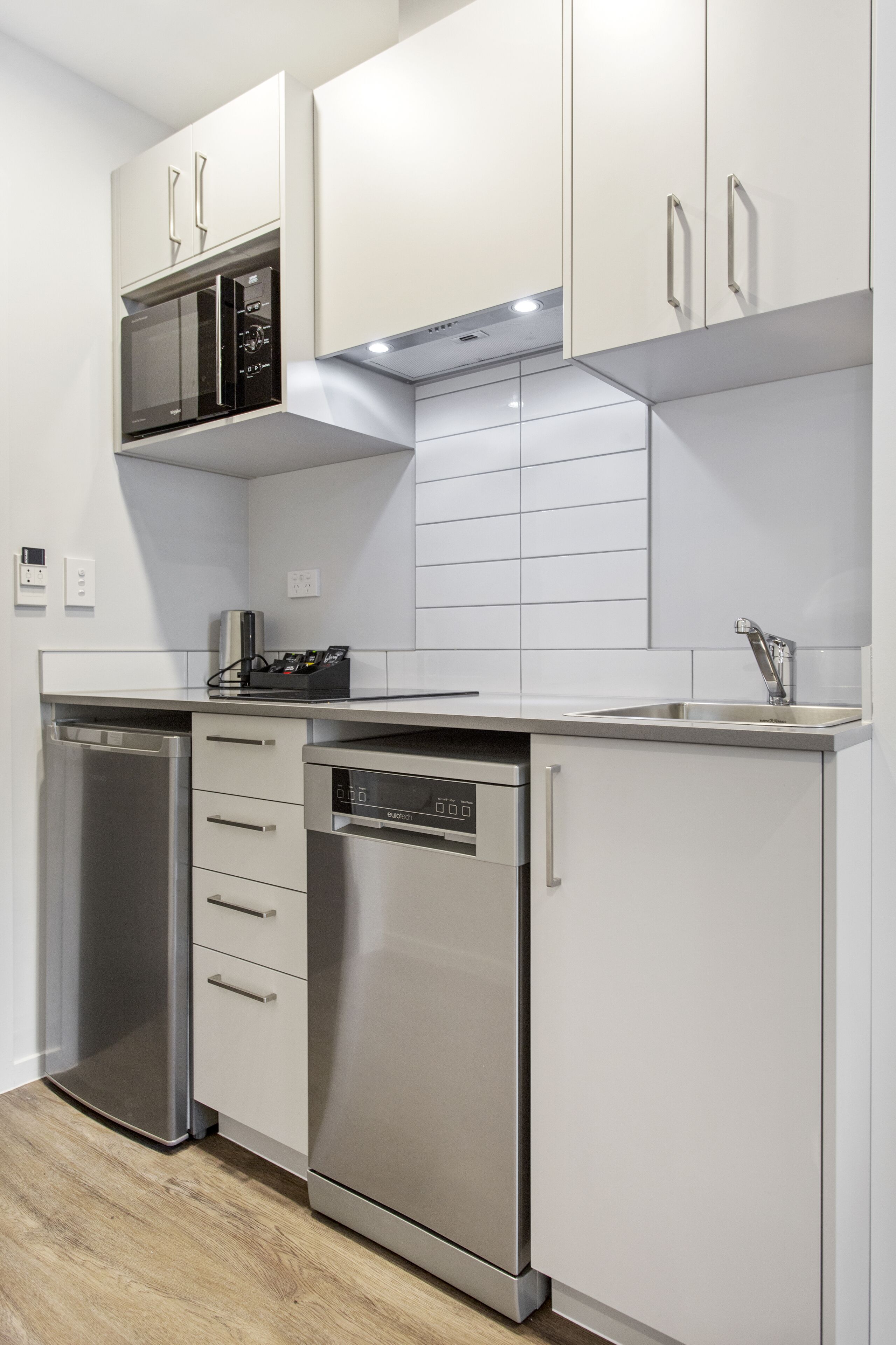 Een compacte keuken met ingebouwde apparatuur, waaronder een vaatwasser, oven en magnetron, met witte kastjes en een betegelde spatwand.