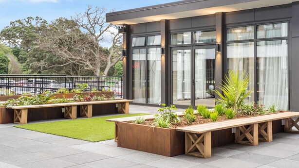 An outdoor seating area with wooden benches and planters, a grassy lawn, and a modern building with large windows and a balcony. - Ramada by Wyndham Newmarket Auckland (Auckland)