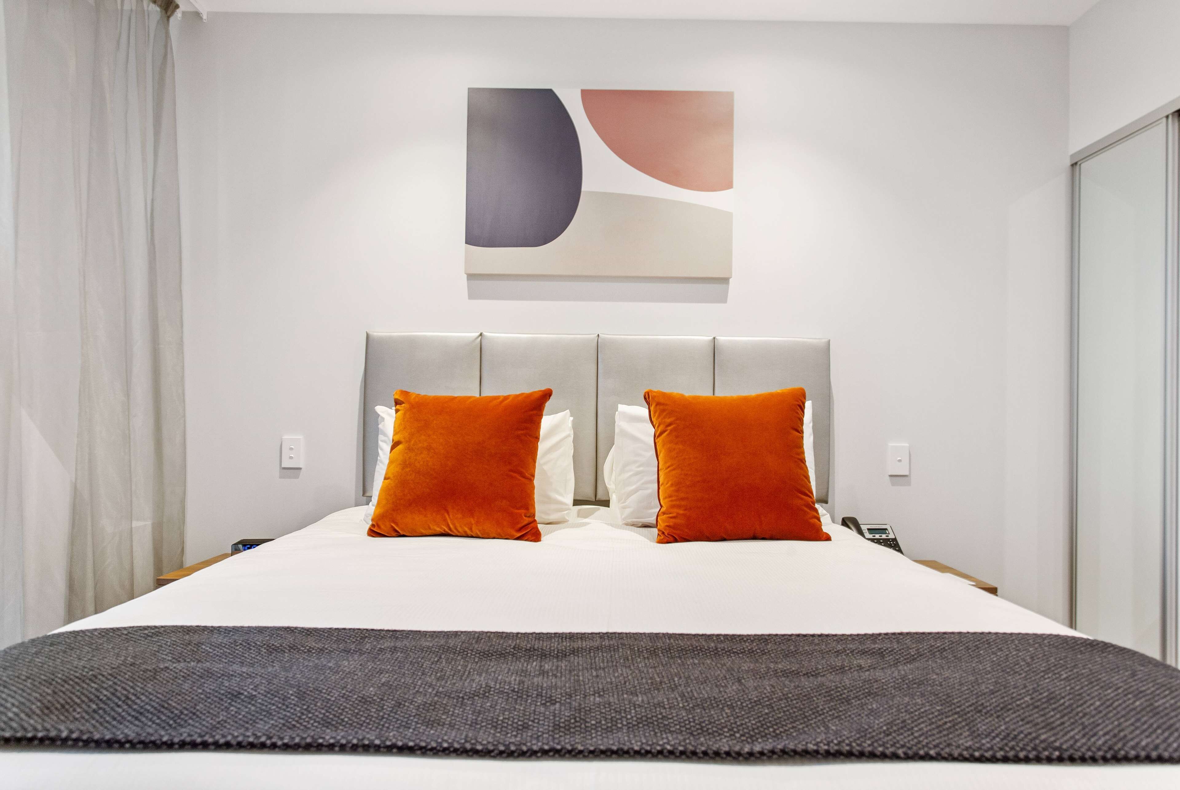 A modern hotel room with a large bed, two orange pillows, a grey headboard, and a piece of abstract art on the wall.