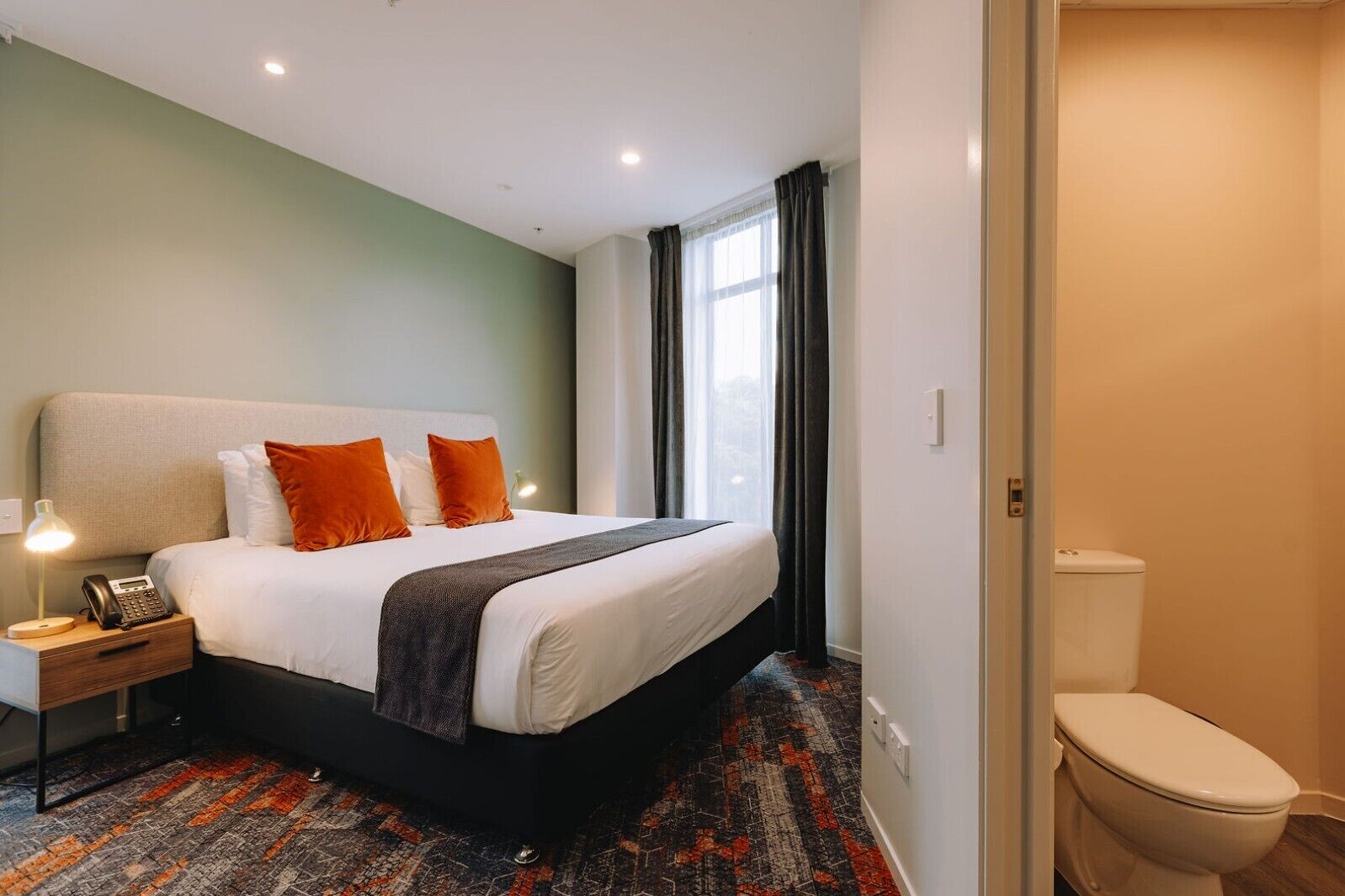 A hotel room with a bed, bedside lamp, telephone, and a view of the outside through a window.