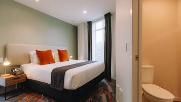 A hotel room with a bed, bedside lamp, telephone, and a view of the outside through a window.