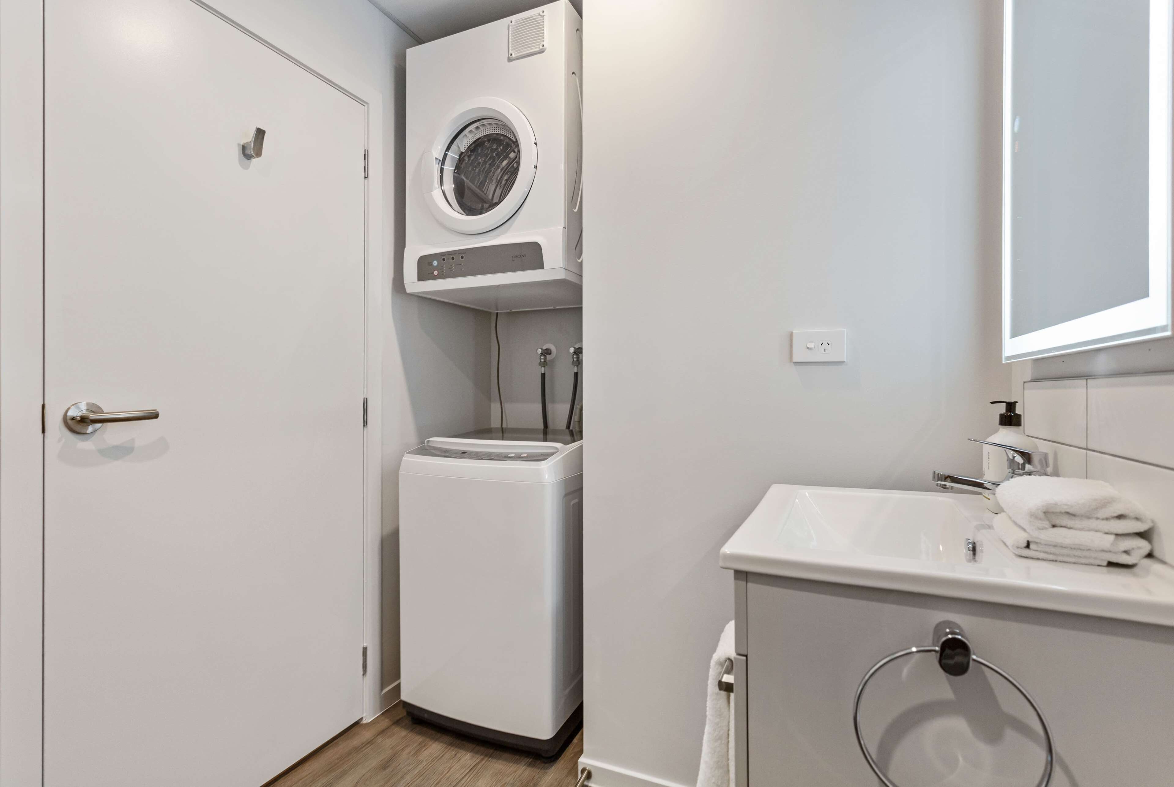 A compact, modern bathroom with a built-in washer and dryer, a sink with a towel rack, and a mirror above the sink.